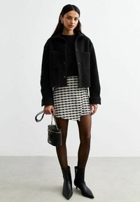 Zwarte, korte fleece jas met twee voorzakken, gecombineerd met een zwart-wit geblokte minirok, zwarte panty en spitsvormige enkellaarsjes.