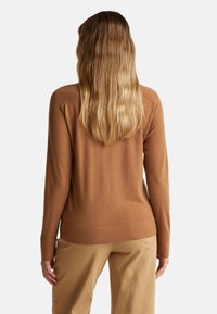 Maglione marrone con maniche lunghe, texture a coste e vita aderente. Indossato con pantaloni beige chiaro, visto da dietro.