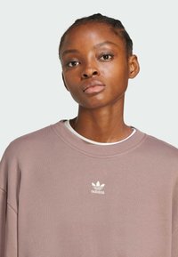 Felpa grigio-rosa con collo rotondo, polsini a coste e un discreto logo Adidas bianco sul petto. Texture liscia e vestibilità comoda.