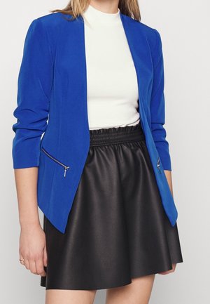Blazer azul ajustado con mangas largas y un bolsillo con cremallera, combinado con una falda de piel sintética negra. Textura suave, diseño a medida.