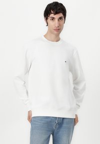 White sweatshirt made of cotton, featuring long sleeves and a round neck. Small logo on the chest; paired with light blue jeans.