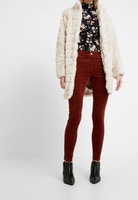 Vadderad krämfärgad faux fur-kappa med ett strukturerat mönster, bärs över en svart blommig långärmad topp och vinröda smala corduroybyxor, kombinerat med svarta ankelstövlar.