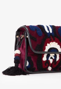 Borsa in velluto con base bordeaux, caratterizzata da motivi in peluche colorati blu, neri e bianchi, e rifiniture in pelle nera. Include un pon-pon.