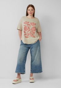 Beżowa, oversize'owa koszulka z kwiatowym wzorem w kolorze koralowym, w połączeniu z jasnoniebieskimi, szerokimi dżinsami i beżowymi sandałami na platformie.