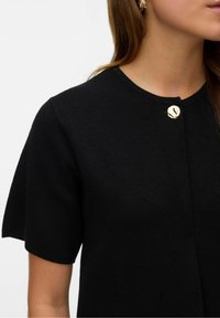 Kobieta ubrana w czarną bluzkę z krótkim rękawem i okrągłym dekoltem, z pojedynczym złotym okrągłym guzikiem blisko kołnierza.