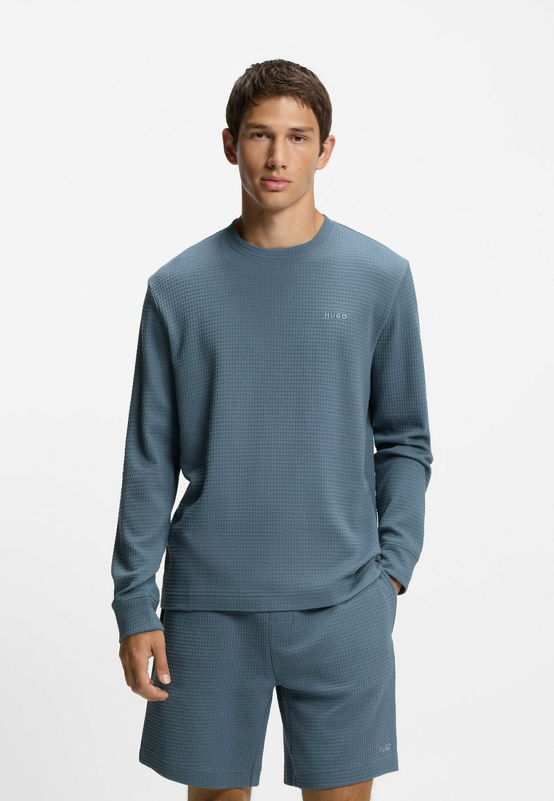 Pitkähihainen sininen pähkinäkakkuinen paita pyöreällä kaula-aukolla ja logolla. Yhdistetty samansävyisten sinisten shortseihin. Sileä kangas ja rento istuvuus.