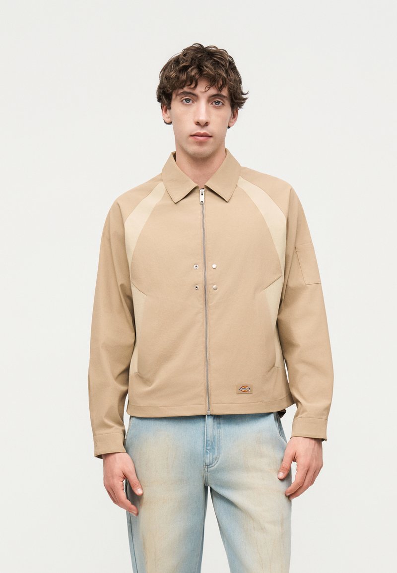 Junger Mann steht und trägt eine beige Jacke mit Reißverschluss mit hellen Einsätzen und hellblauen ausgewaschenen Jeans vor einem schlichten Hintergrund.