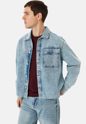 Giacca di jeans blu chiaro con colletto, botttoni frontali e taschino sul petto. Indossata sopra una camicia bordeaux, con una vestibilità morbida e una texture.