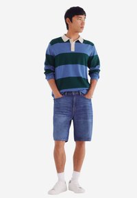 Bunt gestreifter Pullover in Grün und Blau, kombiniert mit Denim-Shorts. Weißer Kragen, Knöpfe am Ausschnitt. Weiße Sneaker und Socken.