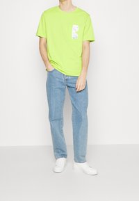 Homme portant un T-shirt à manches courtes vert lime avec un détail de poche brillante, un jean bleu clair et des baskets blanches, debout avec une main dans la poche.