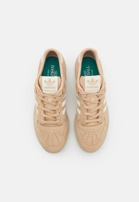 Béžové zamatové športové topánky s bielymi pruhmi, okrúhlou špičkou a perforovanými detailmi. Zelená stielka s označením "adidas Basketball".