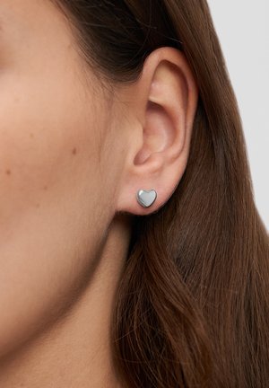Boucles d'oreilles clous en forme de cœur avec une finition argentée brillante, placées sur le lobe, soulignant les contours naturels de l'oreille.