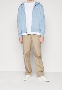 Ljusblå zip-up hoodie med två framfickor, vit t-shirt under och beige cargo-byxor; vita sneakers med strukturerad sula.