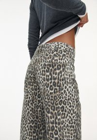 Pantalones de mezclilla con estampado de leopardo en un fondo gris y manchas negras, con dos bolsillos traseros y una cinturilla ajustada. La parte inferior de la camisa es visible por encima.