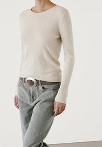 Beige geribbelde top met lange mouwen gecombineerd met grijze wijde jeans en een bruine riem met een grote zilveren gesp. De algehele uitstraling is casual.