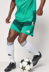 Πράσινες αθλητικές βερμίδες με υφασμάτινο σχέδιο, συνδυασμένες με μαύρα ποδοσφαιρικά παπούτσια και λευκές κάλτσες με μαύρες ρίγες. Ένα μαύρο και λευκό ποδοσφαιρικό μπάλα είναι κοντά.