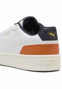 Hvid atletisk sneakers med en tekstureret orange og marineblå hælaccents, med guldfarvede logo detaljer, flade snørebånd og en tyk hvid sål.