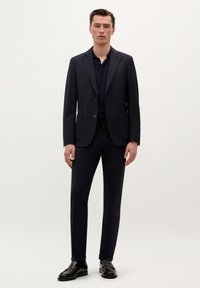 Traje marino con chaqueta de un solo pecho, solapas muesca y dos bolsillos delanteros, combinado con pantalones a juego y zapatos de cuero negro.