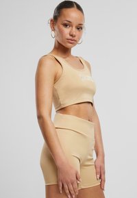 Tan sports crop top og høytlivede shorts laget av glatt stoff, med et frontlogo i hvitt. Outfitten er designet for atletisk bruk.