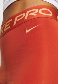 Legíny Nike Pro v jasně červené barvě, s širokým texturovaným pasem, na kterém je logo "Nike Pro" a malý akcent s výšivkou swoosh.