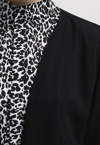 Černý blazer s límečkem a pod ním černobílá košile s potiskem zvířat. Tkanina je hladká a má strukturovaný design.