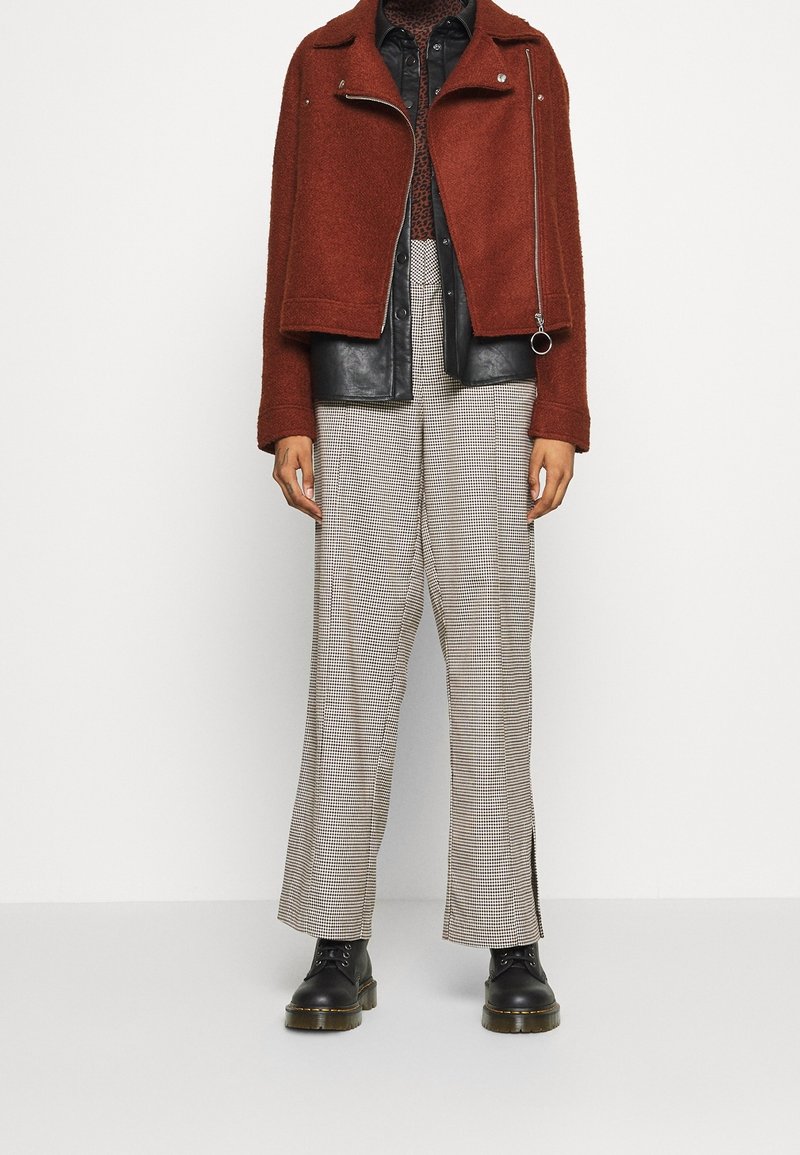 Giacca in misto lana marrone con dettagli in pelle nera, pantaloni a scacchi a vita alta e stivali neri con lacci; dettagli hardware minimi sulla giacca.