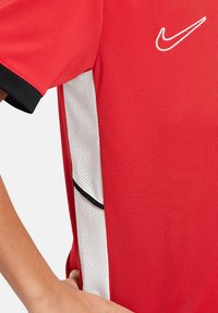 Maglia sportiva rossa con maniche corte, bordature nere e pannelli in rete bianca con piccoli fori. Logo ricamato sul davanti.