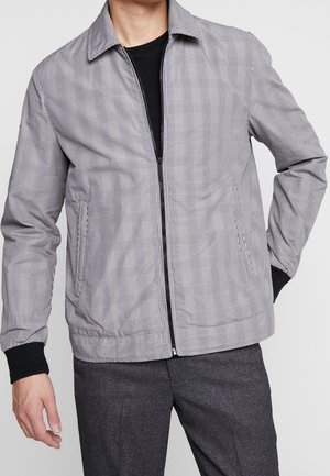 Hombre vestido con chaqueta gris a cuadros con cremallera sobre camiseta negra y pantalones gris oscuro, de pie con una mano en el bolsillo y la otra relajada.