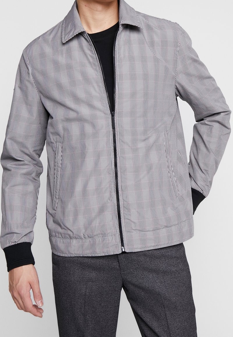 Homme portant une veste zippée à carreaux gris par-dessus une chemise noire et un pantalon gris foncé, debout avec une main dans la poche et l'autre détendue.
