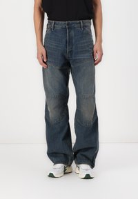 Jean en denim avec une coupe décontractée, affichant une couleur bleue délavée, un design en patchwork et des détails de genoux renforcés. Portés avec des baskets blanches.