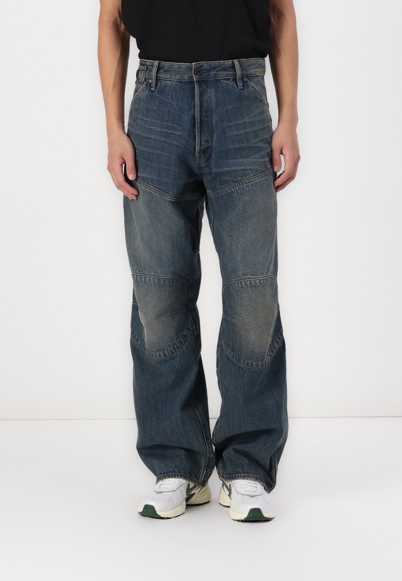 Jean en denim avec une coupe décontractée, affichant une couleur bleue délavée, un design en patchwork et des détails de genoux renforcés. Portés avec des baskets blanches.