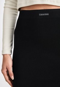 Fekete bordázott kötött szoknya Calvin Klein logóval a derékrészen, világos színű, testhezálló, hosszú ujjú felsővel párosítva. A texturált anyag látható.