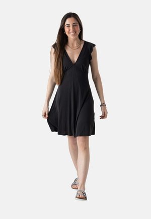 Mujer caminando hacia adelante sonriendo, vistiendo un vestido negro sin mangas y sandalias plateadas, sosteniendo el dobladillo del vestido con una mano.