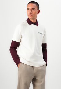Polo blanc à manches courtes avec un col bordeaux, arborant un logo vert sur la poitrine et de longues manches bordeaux en dessous. Pantalon de couleur neutre.