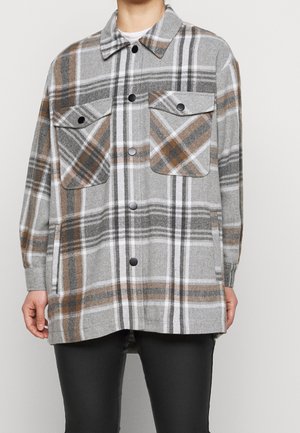 Persona con chaqueta camisa de cuadros gris, marrón y blanco con bolsillos en el pecho y pantalones negros, de pie contra un fondo liso.