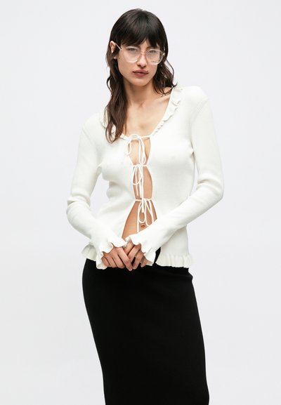 Άσπρο πουλόβερ με ριγέ υφή, βαθύ V-λαιμόκοψη με δέσιμο, λεπτομέρεια με βολάν κατά μήκος της λαιμόκοψης και των μανικιών, συνδυασμένο με μαύρη εφαρμοστή φούστα.