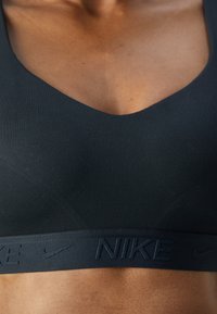 Svart sportsbh med V-halsdesign, laget av glatt stoff med en bred kant som har preget "NIKE"-logo.
