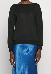 Haut noir à manches longues avec un large décolleté, en matière pailletée. Porté par-dessus une jupe satin bleu clair avec une texture froissée.