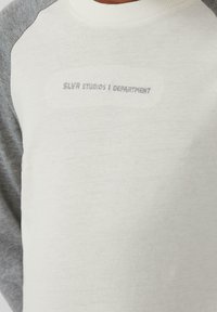 Weißes und graues Langarmshirt mit Raglanärmeln und gesticktem Text "SLVR STUDIOS | DEPARTMENT" auf der Vorderseite. Weicher Stoff.