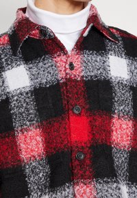 Close-up af en rød, sort og hvid ternet uldskjorte med knapper, lagdelt over en hvid rullekravetrøje på en person.