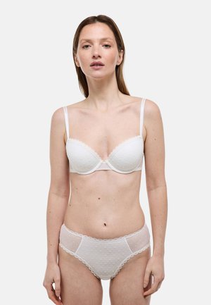 Femme à la peau claire et aux cheveux bruns portant un soutien-gorge blanc texturé et une culotte en maille transparente assortie, debout devant un fond uni.