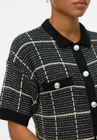 Polo shirt tricoté à motifs noir et blanc, avec un col côtelé, des manches courtes, des accents de boutons en argent et un détail de poche.