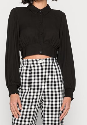 Kobieta ubrana w czarną krótko skrojoną koszulę na guziki z długimi rękawami zakończonymi marszczonymi mankietami oraz w czarno-białe spodnie w kratę o wysokim stanie.