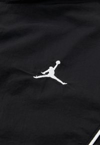 Black fabric with a smooth texture featuring a white embroidered Jumpman logo in mid-air pose, capturing motion and athleticism.