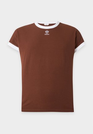 Un jeune homme se tient debout, portant un T-shirt marron avec une bordure blanche, un pantalon rayé multicolore et des chaussures foncées avec des bouts dorés sur un fond uni.