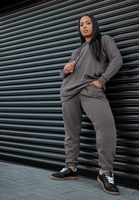 Grijze geribbelde hoodie en bijpassende trainingsbroek met zakken, gecombineerd met zwarte sneakers met witte veters en rubberen zolen, tegen een zwarte achtergrond.