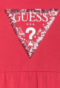 Czerwona bawełniana koszulka z okrągłym dekoltem, ozdobiona trójkątną łatą z cekinów z napisem "GUESS U.S.A." oraz centralnym znakiem zapytania, poniżej zebranym detalem na dole.