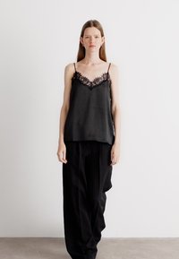 Svart satin cami med tynne stropper og blonderkant ved utringningen, kombinert med løse svarte bukser. Jevn tekstur, minimalistisk design.