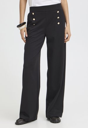 Pantalon noir à jambes larges avec six boutons dorés sur le devant, porté avec des chaussures noires et des bracelets au poignet gauche.