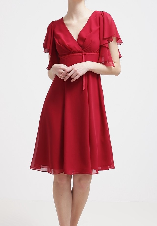 Femme portant une robe rouge foncé longueur genoux avec des manches flottantes superposées et une ceinture nouée à la taille, debout les mains jointes.
